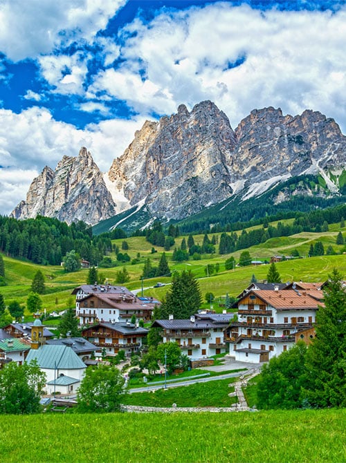A scenic alpine village with wooden houses and green fields nestled at the base of rugged, rocky mountains under a partly cloudy blue sky.