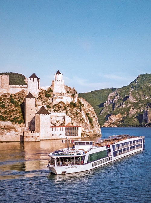 A river cruise ship sailing along the Danube past a medieval stone fortress perched on rocky cliffs, with green hills rising on either side under a clear blue sky.