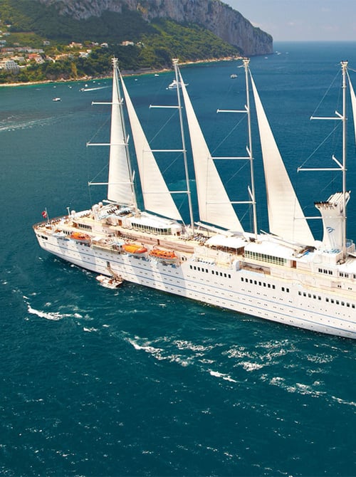 A large white cruise ship with multiple tall sails traveling through deep blue coastal waters, with a green hillside and seaside town in the background.