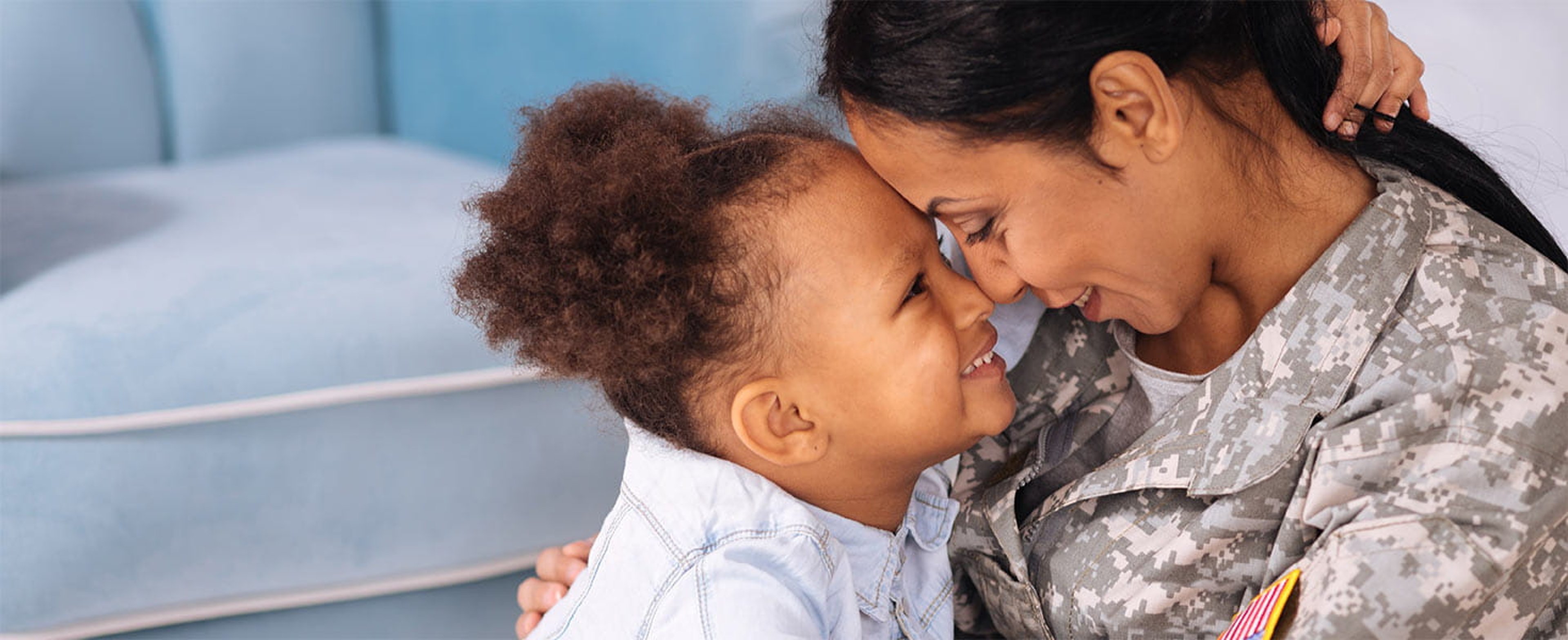 A military mom in uniform lovingly embraces her young daughter, their heads touching eye to eye conveying deep emotion and connection.