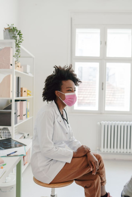 A healthcare professional wearing a lab coat and a face mask, sitting thoughtfully on a stool.