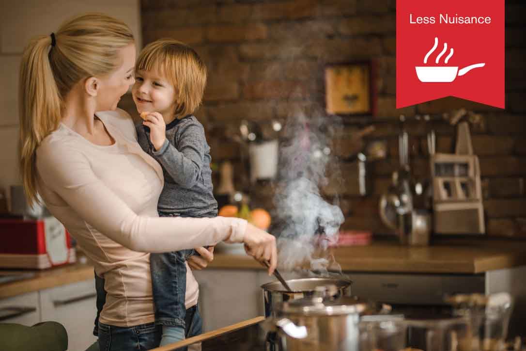 Less Nuisance.  A woman holds her young son in her arms while she cooks over the stovetop.