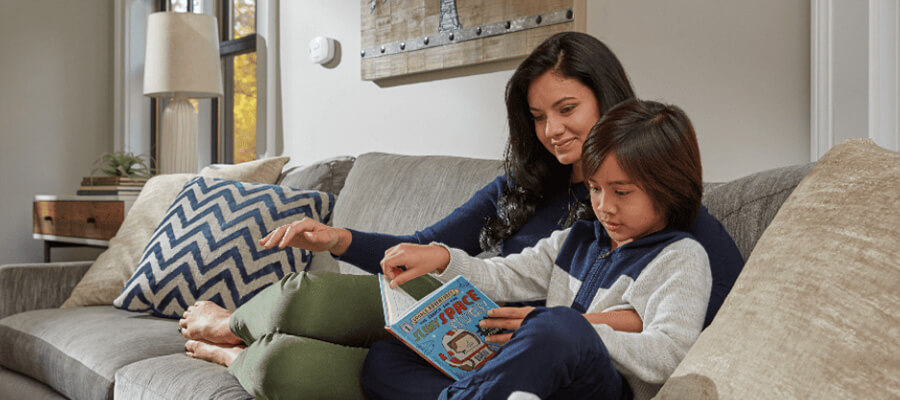 A mother sits on a couch with her young son as he reads a book.