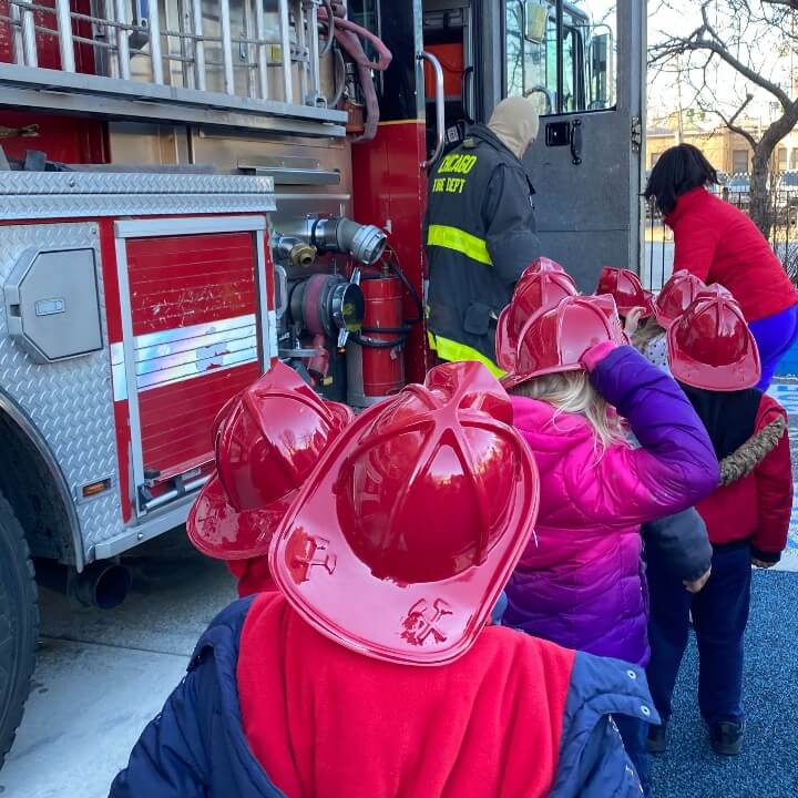 Children wearing firefighter hats line up near a firetruck to receive souvenirs.