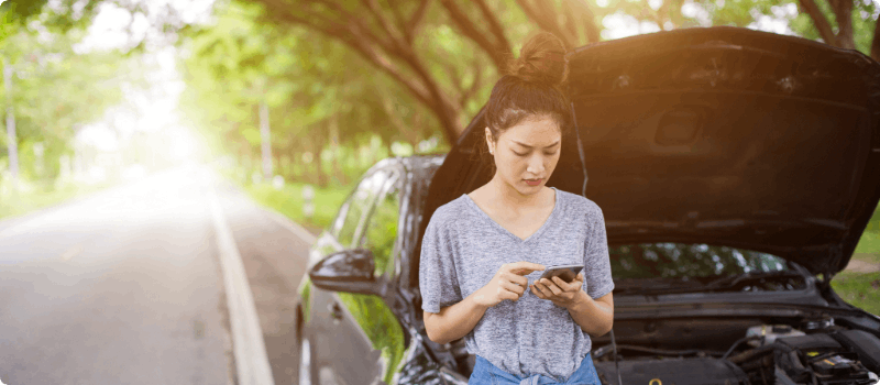 A person standing on the roadside looking at their phone while leaning against their broken-down vehicle.