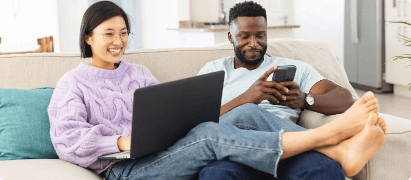 A happy couple resting on their couch while working on their laptop and phone.