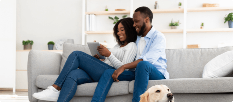 A happy couple sitting on their living room couch and looking at a computer tablet. 