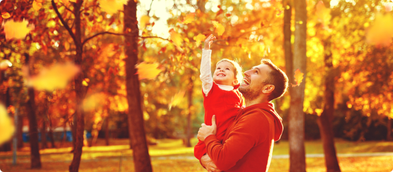 A parent holding their child as they enjoy the surrounding trees' fall leaves.