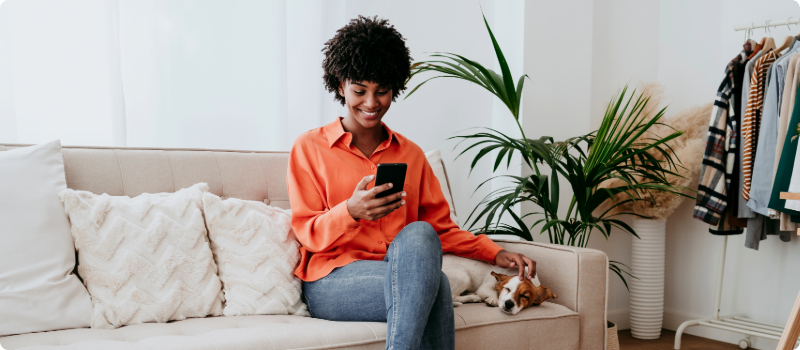 A person sitting on their couch and petting their dog while looking at their phone. 