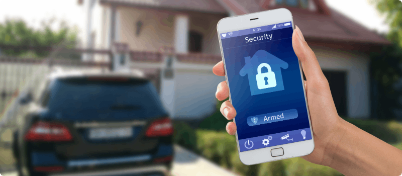 Person holding a smartphone displaying their home security app in front of their home.