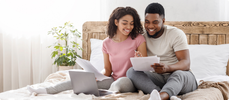 A happy couple sitting on their bed, reviewing documents with an open laptop in front of them. 