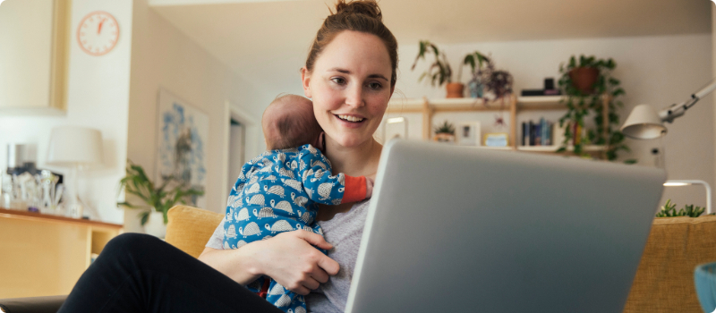 A mother holding her infant at a table while working on her laptop.