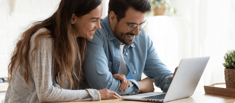 A smiling couple sitting at a desk while looking at their laptop.