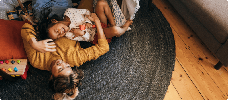 A parent cuddling and laughing with their child while lying on the floor.