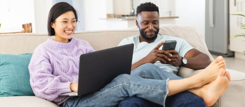 A couple sitting on their couch, looking at their phone and laptop.