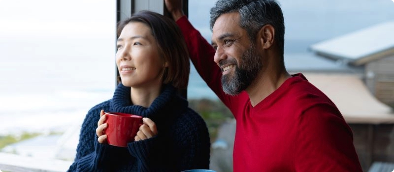 A smiling couple standing by a window while looking out at the ocean.