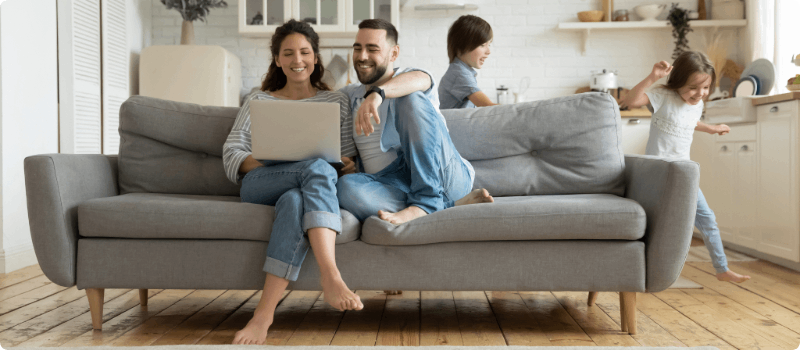 Parents sitting on their living room couch in front of a laptop as their kids play in the background.