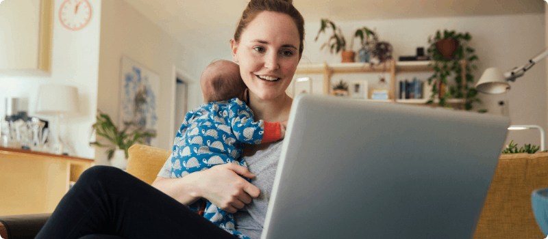 A new parent holding her infant while looking at her laptop.