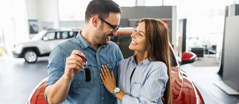 Couple at a car dealership, holding up a set of keys.  