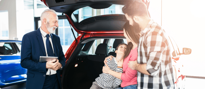 Family of three sitting in front of their new car while the salesperson holds the contract. 