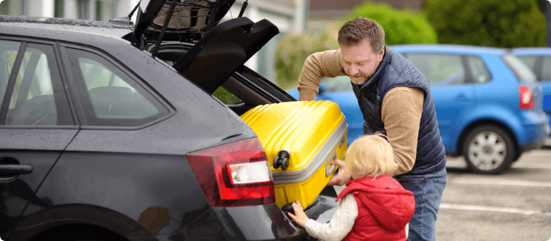 Parent and child pulling luggage from their vehicle's trunk in a hotel parking lot.