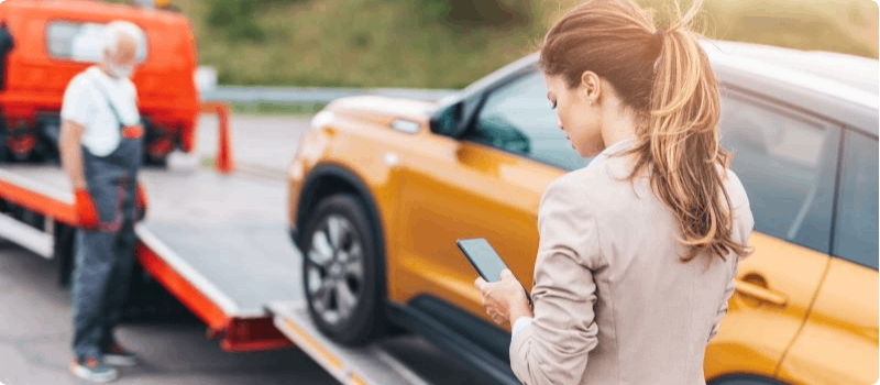 Woman looks at her smartphone while roadside assistance loads her vehicle onto the back of their truck.