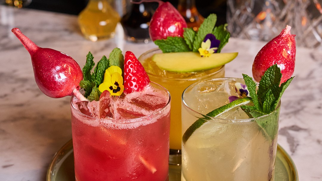 Tray of three colorful signature cocktails in short glasses, garnished with sugared strawberries, mint, and flowers.