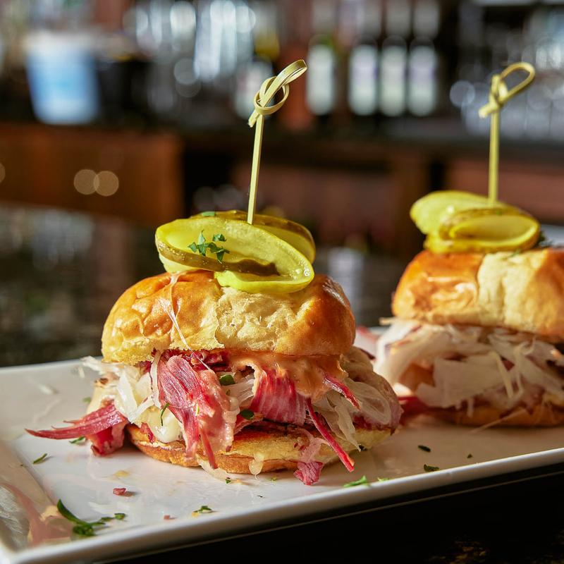 Two corned beef sliders with pickle garnish on white plate.