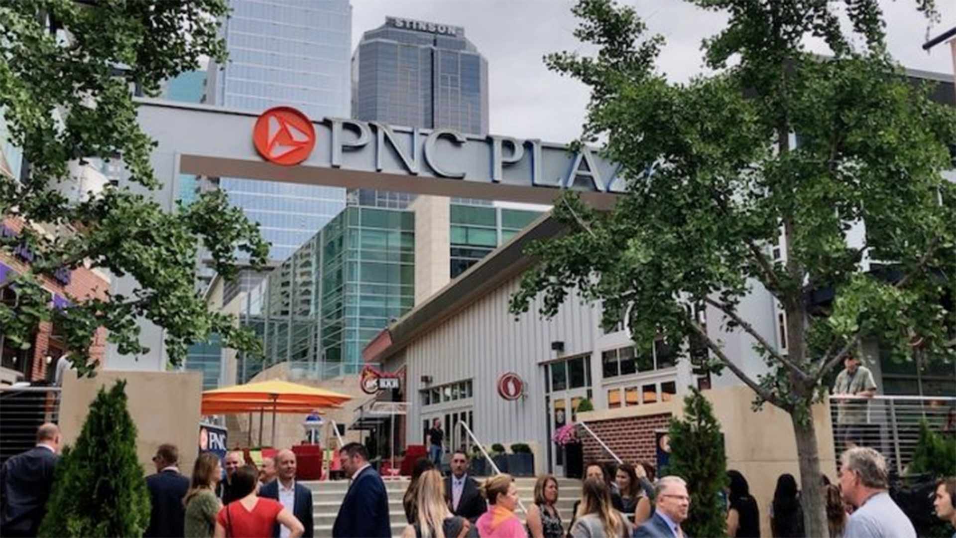 The entrance to PNC Plaza on a sunny afternoon is pictured with patrons ascending the stairs between two green trees. 