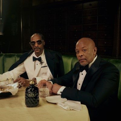 Snoop Dogg and Dr. Dre sitting in a green banquet booth with Still G.I.N. bottle on the table in front of them.