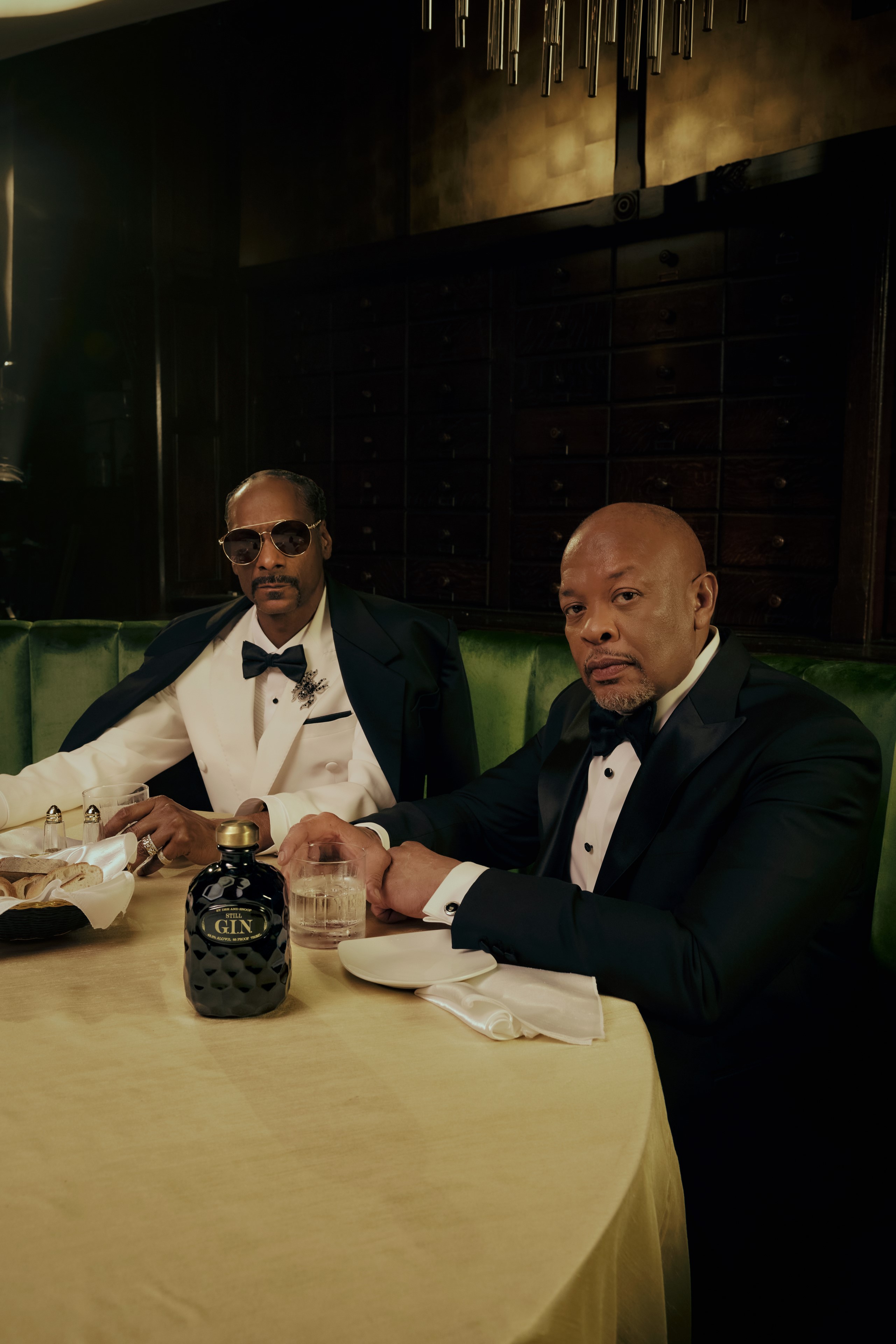 Snoop Dogg and Dr. Dre dressed in formal wear sitting in a booth with Still G.I.N. bottle on table.