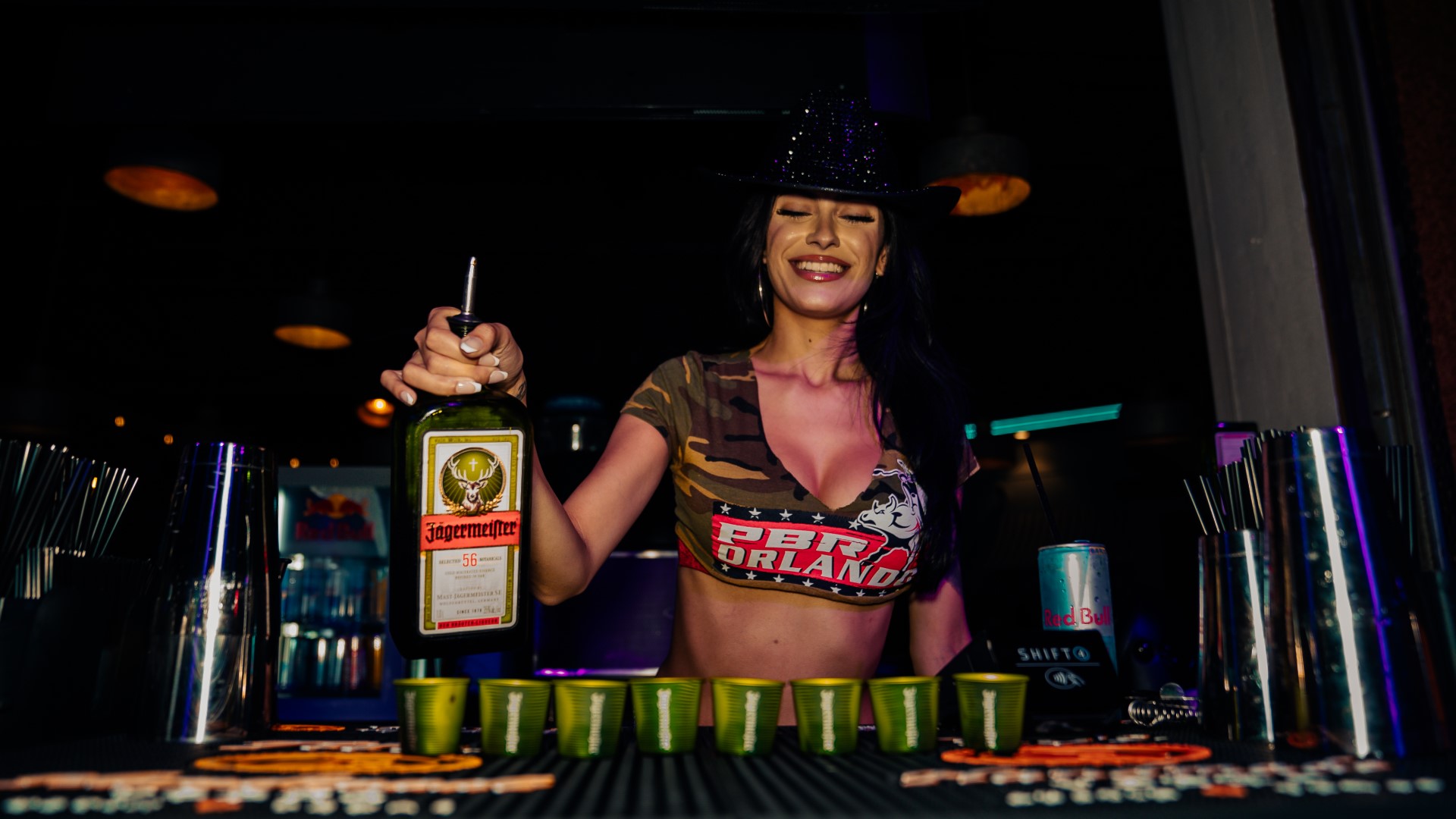 Female bartender behind the bar of PBR Orlando wearing black cowboy hat and cropped top pouring a line of shots.
