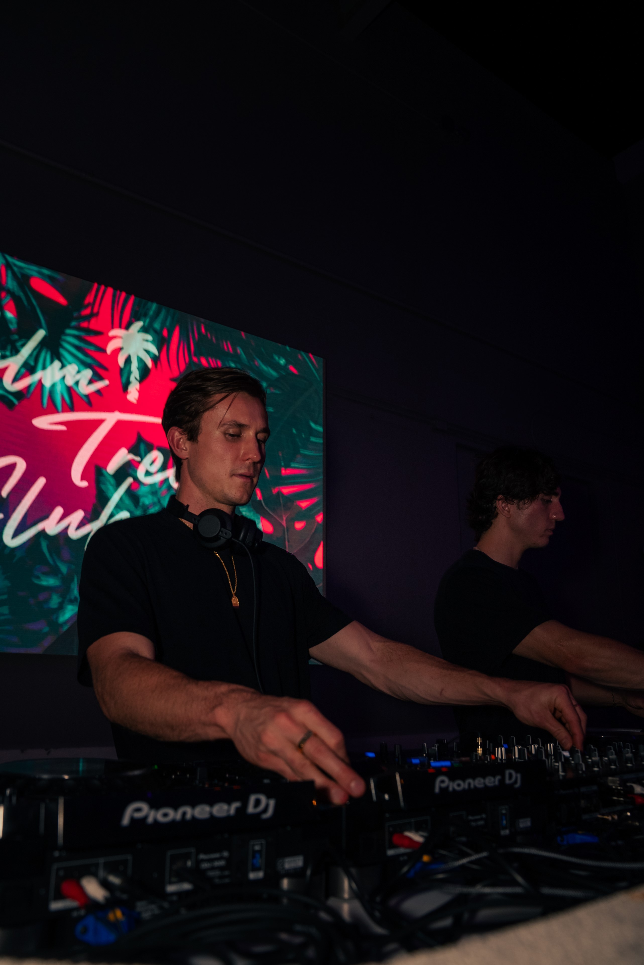 Djs at turntables with Pink and green Palm Tree Club sign behind them.