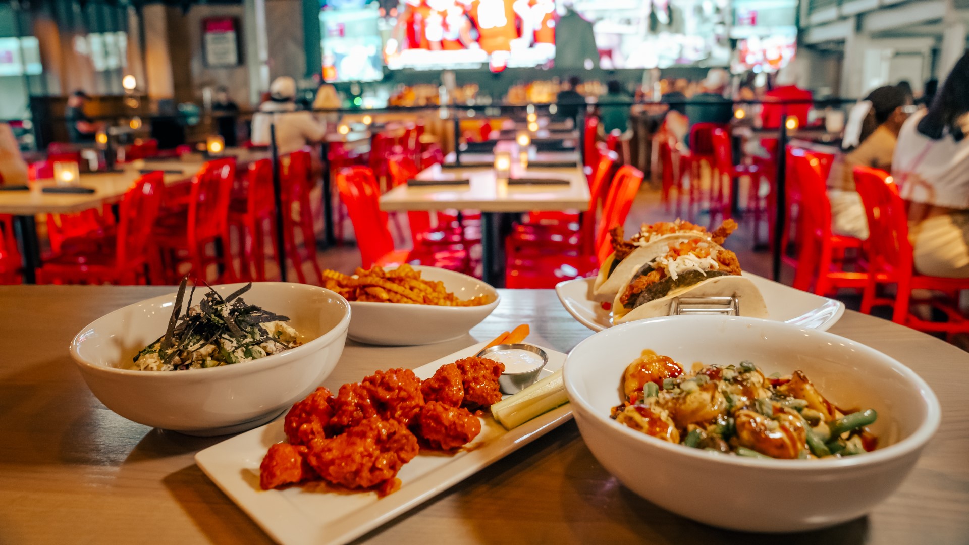Food (chicken wings, fries, tacos) on white plates and bowls on a table in the dining area of Sports & Social at Live! at the Pointe Orlando.