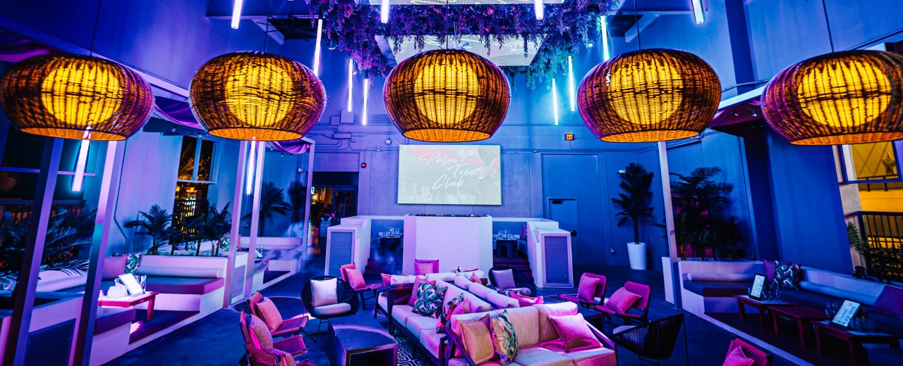 Palm Tree Club lounge seating and DJ booth at night with warm blue and pink lighting and large overhead lantern lights.
