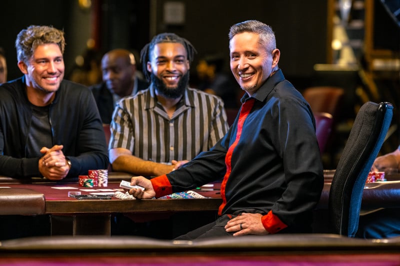 a table games dealer sits at the table, smiling at the camera. players sit behind him