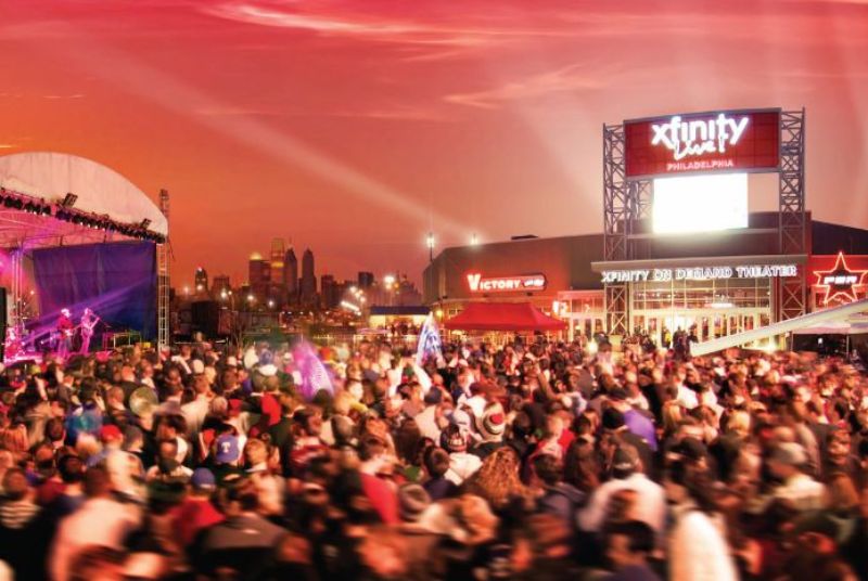 A concert crowd cheers on the band while the surrounding Philly city skyline and Xfinity Live! theater glow from the sunset.