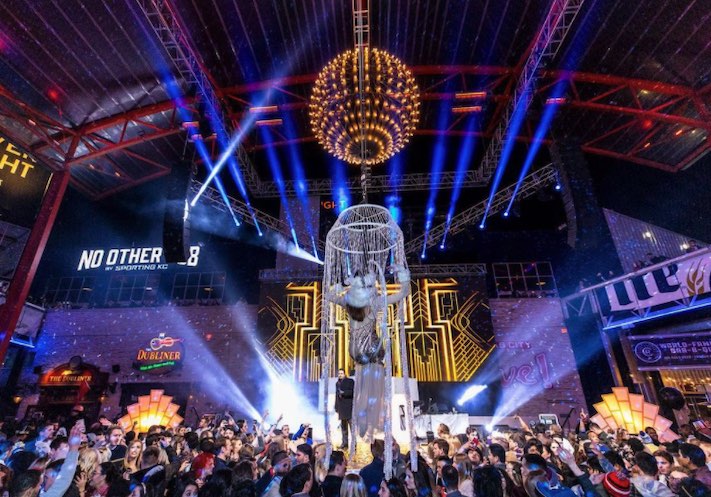 A lighted ball hangs over a glittery-dressed acrobat sitting in a hoop above the crowd as an emcee looks on.