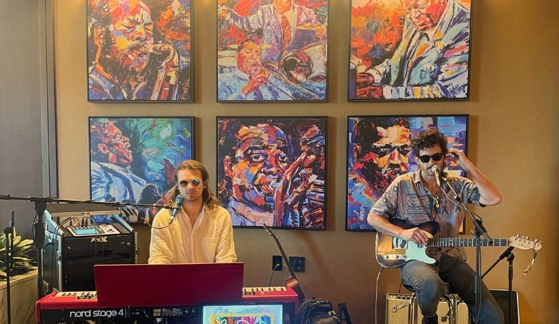 image of two musicians in front of a piano and sitting on a stool with a guitar