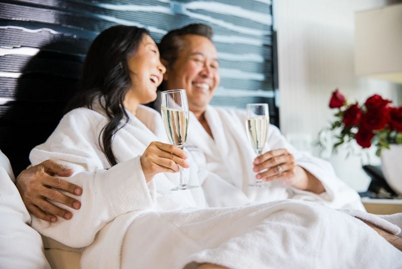 Middle aged Asian couple sitting in bath robes on a bed with glasses of champagne