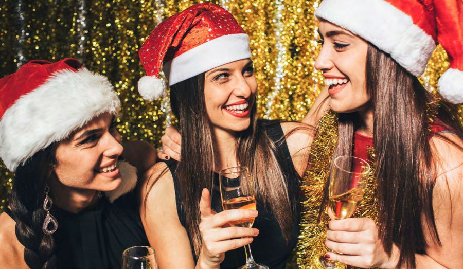 3 women with santa hats on smiling and laughing in front of a glittery gold background