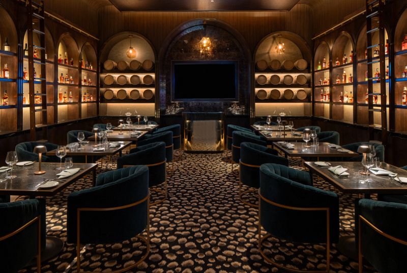 Dim room with shelves stocked with different types of bourbon, dining tables with plush dark blue chairs.