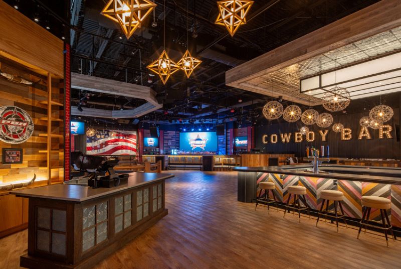 Large room with a check in desk, a bar towards the front of the room, a bar and a stage on the far side of the room, and a VIP booth area with the words Cowboy Bar on the wall in lights.