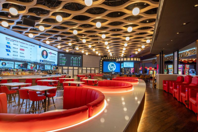 Large sports bar area with different types of booth and tables. All booth seating and regular seating is red, while the tables are white. Large TV screens cover the left side of the room along with a bar lining this area.