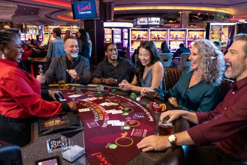 Group of guests looking happy and excited around a blackjack table with dealer and cards and chips. Guests are enjoying cocktails.