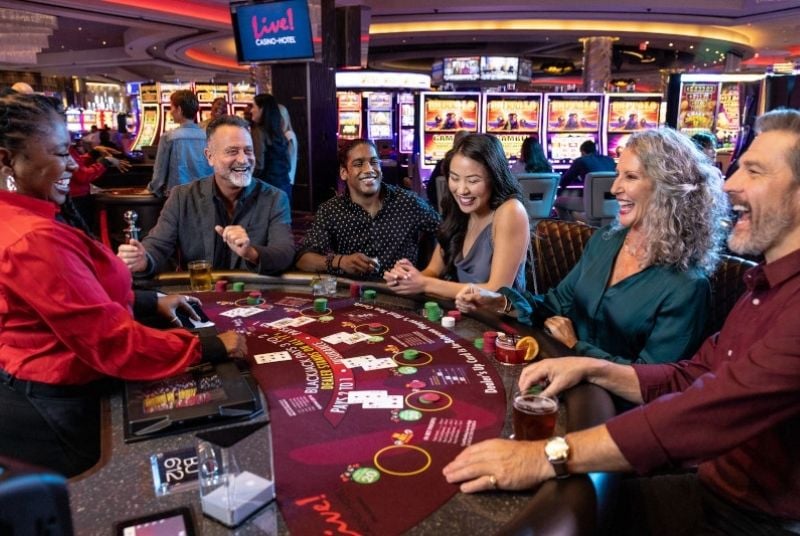 Group of guests looking happy and excited around a blackjack table with dealer and cards and chips. Guests are enjoying cocktails.