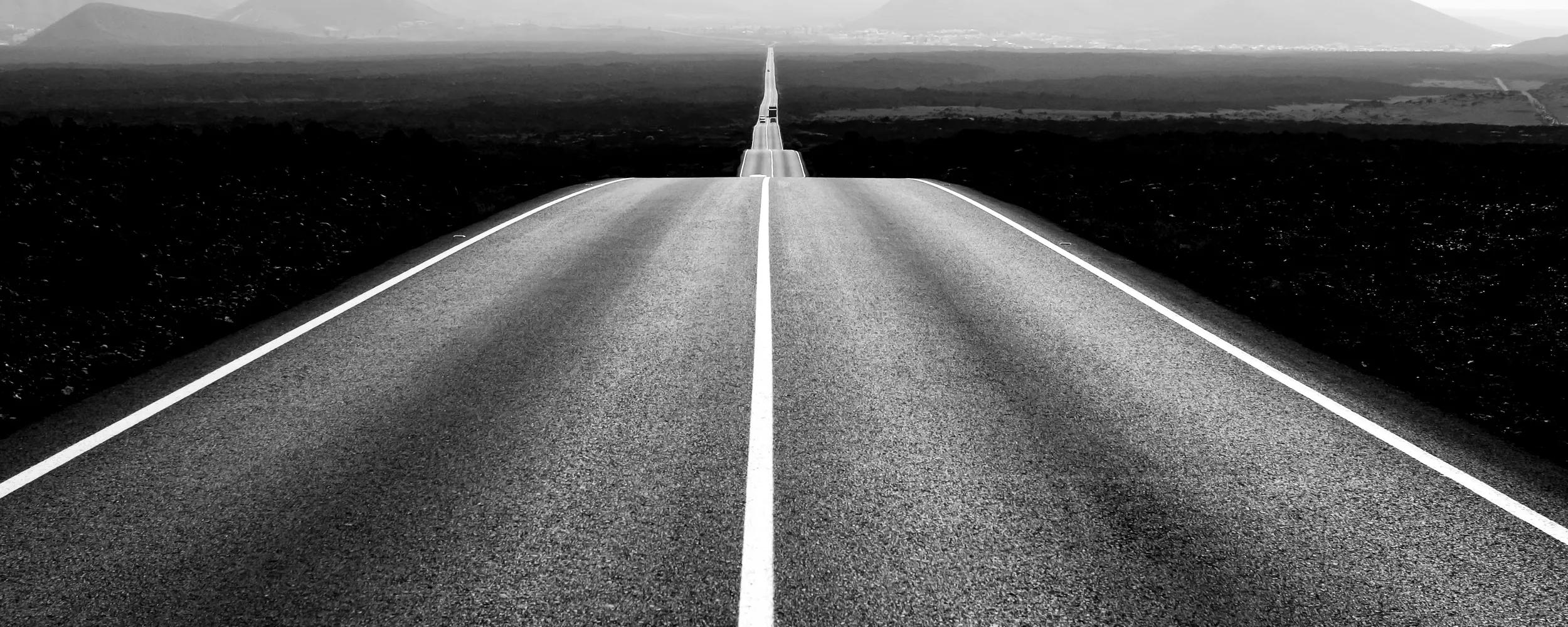 Black and white scene including a long road leading to the foot of the mountains