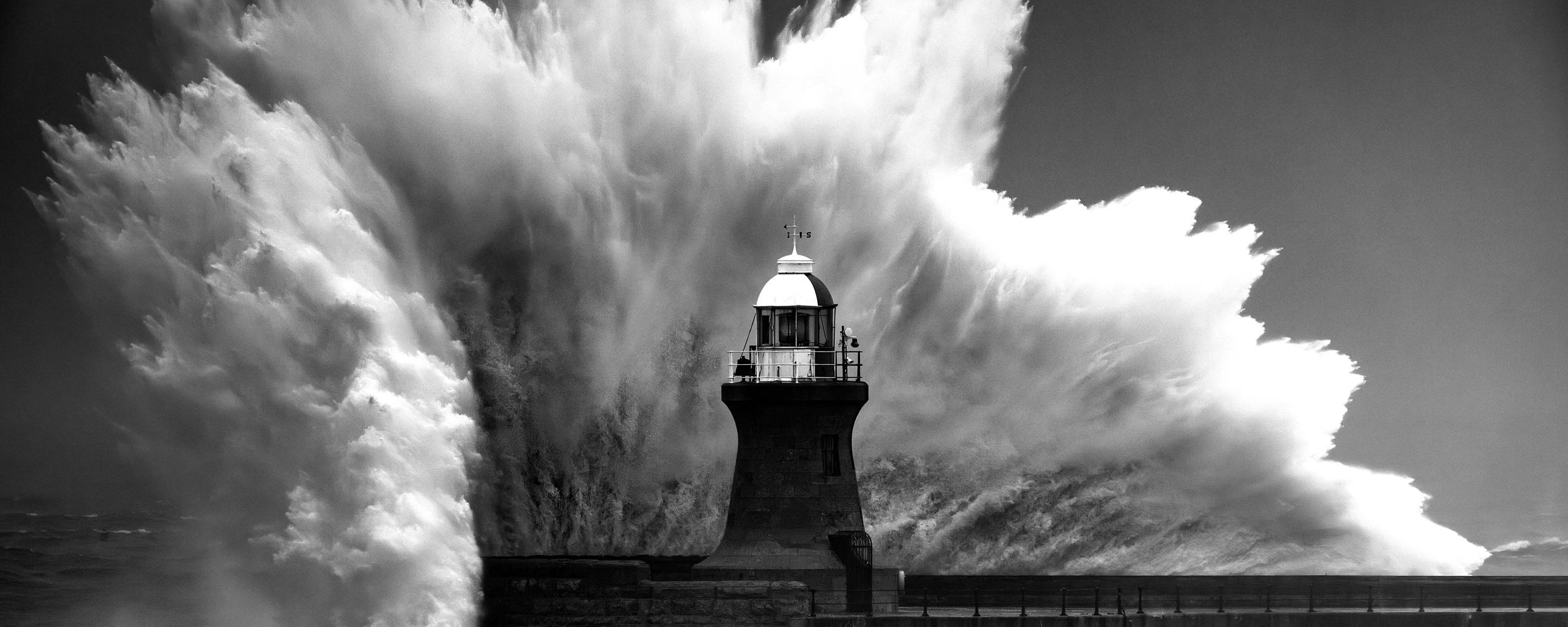 A wave crashing into a pier covering a light house