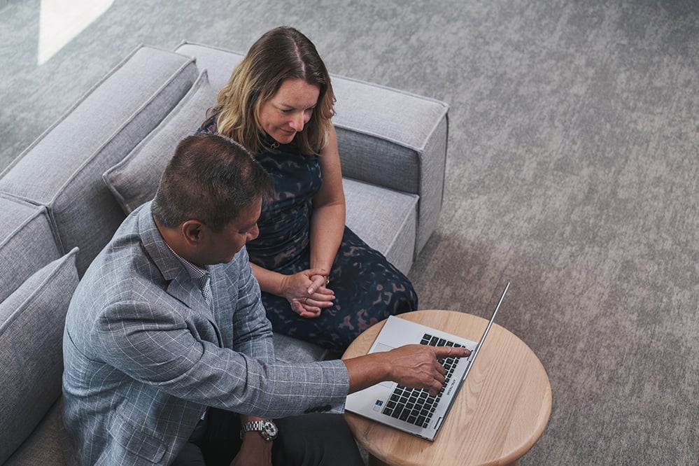 Two professionals sitting on a grey sofa reviewing information on a laptop placed on a small round wooden table, with one person pointing at the screen in a modern office setting.
