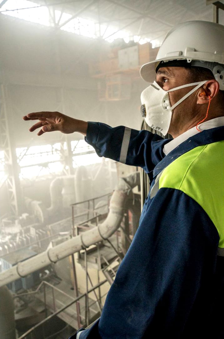 Industrial worker in a white hard hat, safety vest, and face mask pointing at machinery inside a facility
